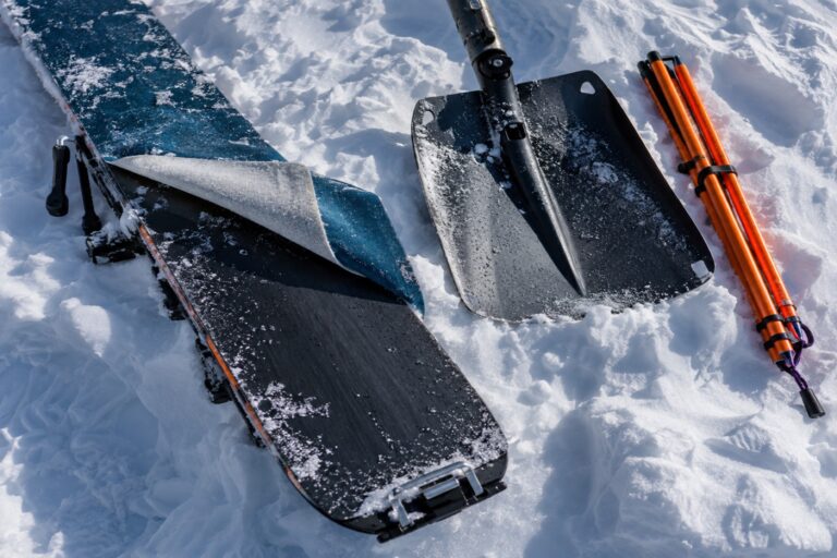 A touring ski with its climbing skin half peeled back lying in the snow, shovel and probe beside it